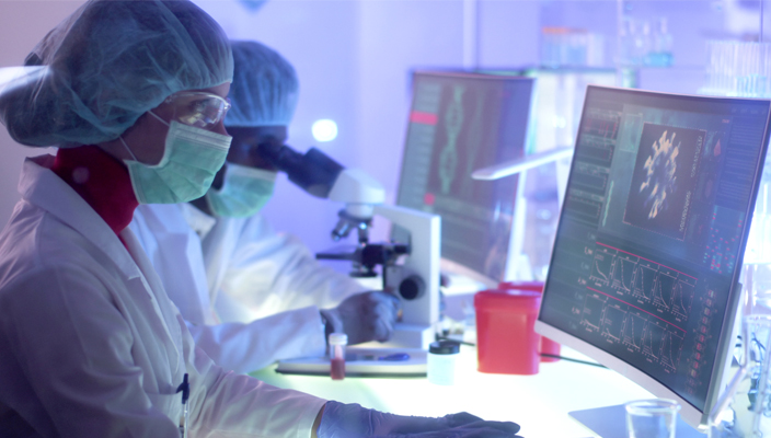 Investigadores científicos en un laboratorio con cubrebocas y vestimenta de protección. Uno mira a través de un microscopio mientras el otro trabaja en una computadora. 