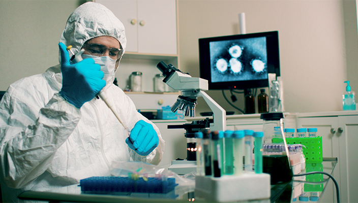 Un investigador científico en un laboratorio, vestido con ropa de protección, cubrebocas y guantes, usa una pipeta para inyectar algo en un tubo de ensayo. A su alrededor hay tubos de ensayo, un microscopio y un monitor de computadora.