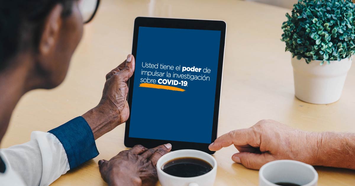 Una persona mira la pantalla de una tableta que muestra un texto que dice, “Usted tiene el poder de impulsar la investigación sobre COVID-19”, mientras la mano de alguien más apunta a la pantalla, junto a una taza de café. 