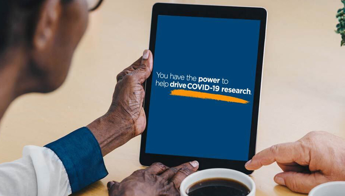 Over a cup of coffee, person looks at a tablet computer displaying the text “You have the power to help drive COVID-19 research,” while someone else’s points to the screen.