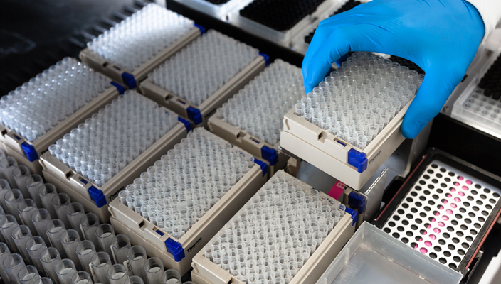 Samples from participant sites in clear test tubes are automatically dispensed into smaller portions and removed for storage by a gloved technician.