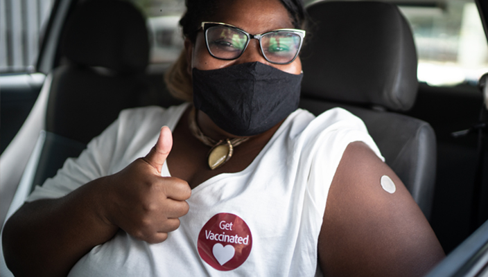 A woman sitting in her car, her sleeve rolled up, wearing a mask and a sticker that reads “Get Vaccinated,” and giving a thumbs up sign.