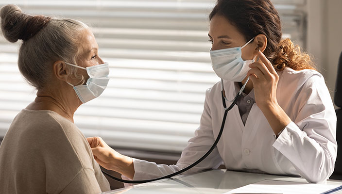 A health care provider using a stethoscope on a patient. Both are wearing masks.