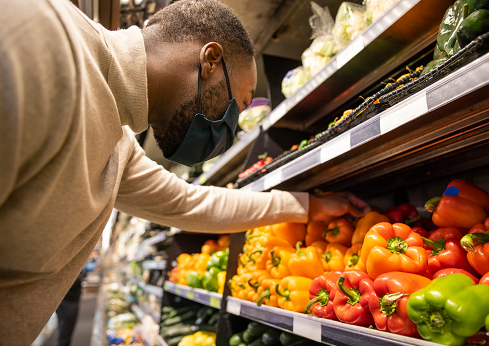 A man wearing a mask selects bell peppers from a shelf in a grocery store produce aisle.
