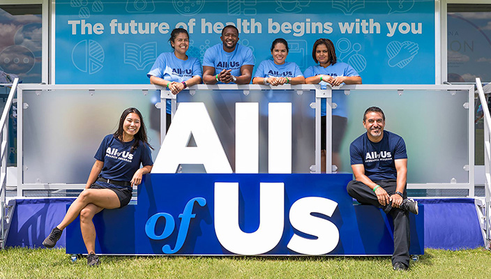 Seis personas sentadas al lado o de pie detrás del logo 3D de All of Us. Detrás de ellos hay una pancarta que dice en inglés “The future of health begins with you” (El futuro de la salud comienza con usted).