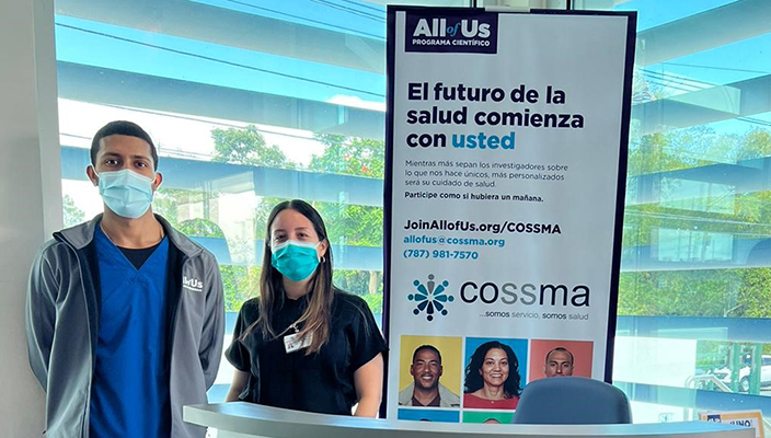 Two people wearing masks standing next to a banner that reads in Spanish, “All of Us: El futuro de la salud comienza con usted” 